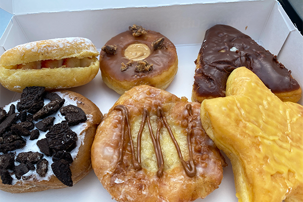 Six donuts in a box. Clockwise from top left: strawberries and cream in a donut, peanut butter cup donut, A-shaped chocolate cream-filled donut, an orange star, a fritter with apple in the middle and a caramel drizzle, and a cookies and cream donut with pieces of dark Oreo cookie | Multiple Locations