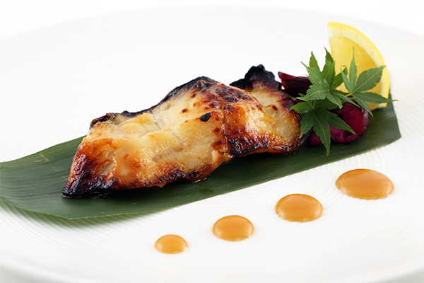 A miso-glazed piece of cod on a green leaf with four dots in the foreground
