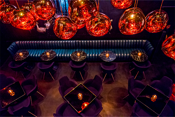 Overhead shot of dark bar with square tables and purple chairs, a booth with purple chairs, and red hanging light fixtures